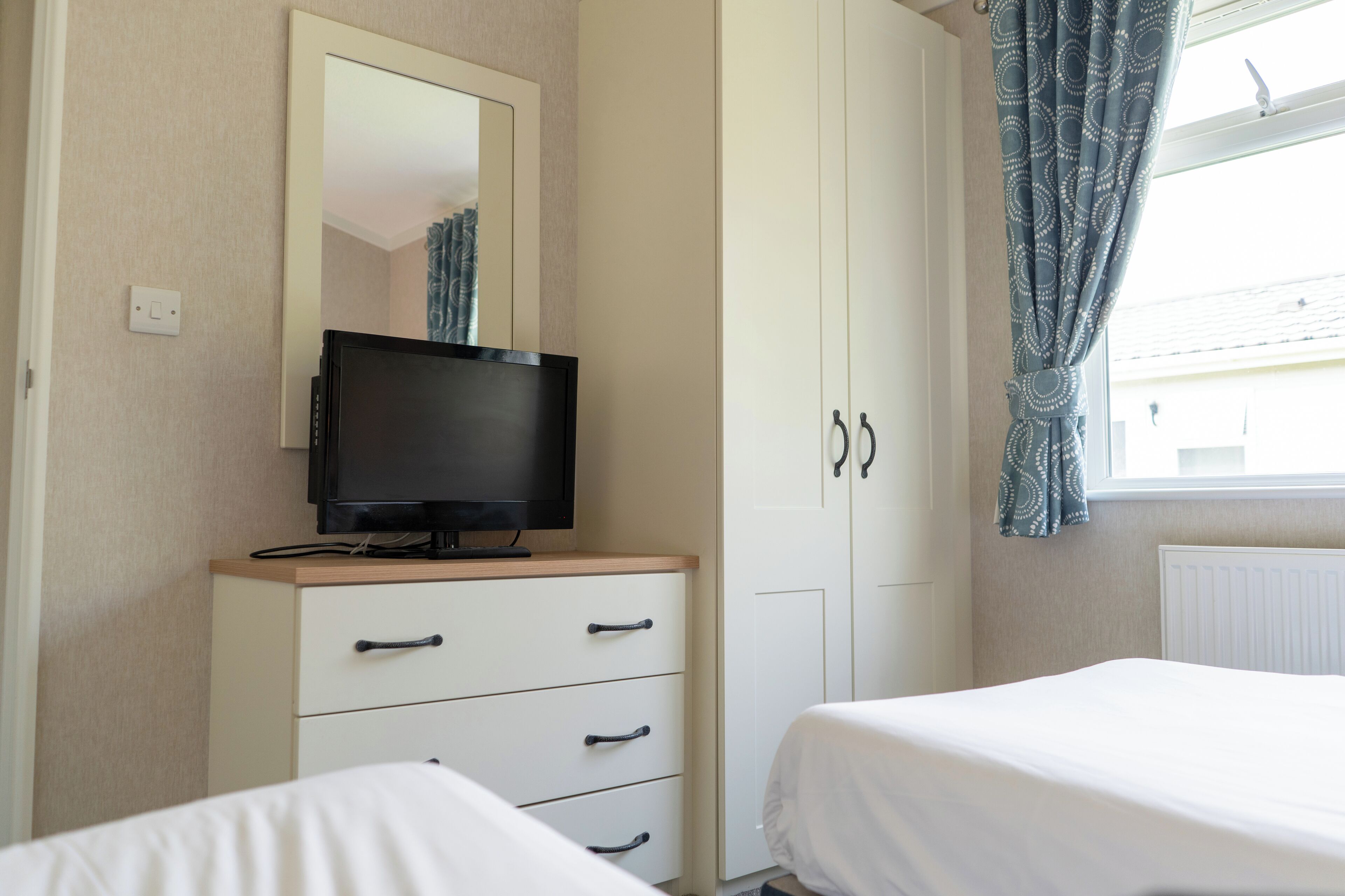 A bedroom with a TV on a dresser, a wardrobe, and a window with curtains.