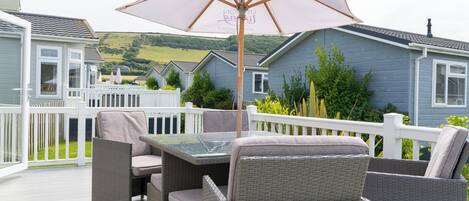 A patio with wicker furniture and a table, surrounded by a white fence, overlooking a green landscape.