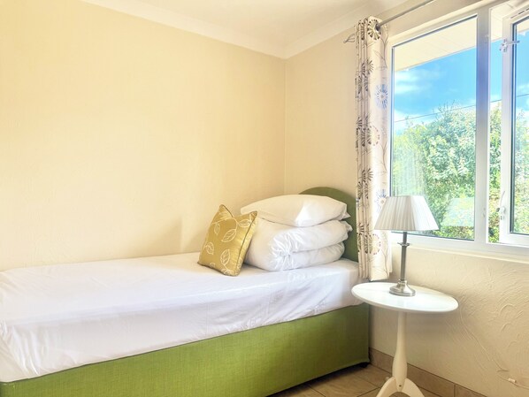 A single bed with a green headboard, white bedding, and a gold-patterned pillow. A small white table with a lamp is beside the bed. - Croyde Bay Holiday Resort (Braunton)