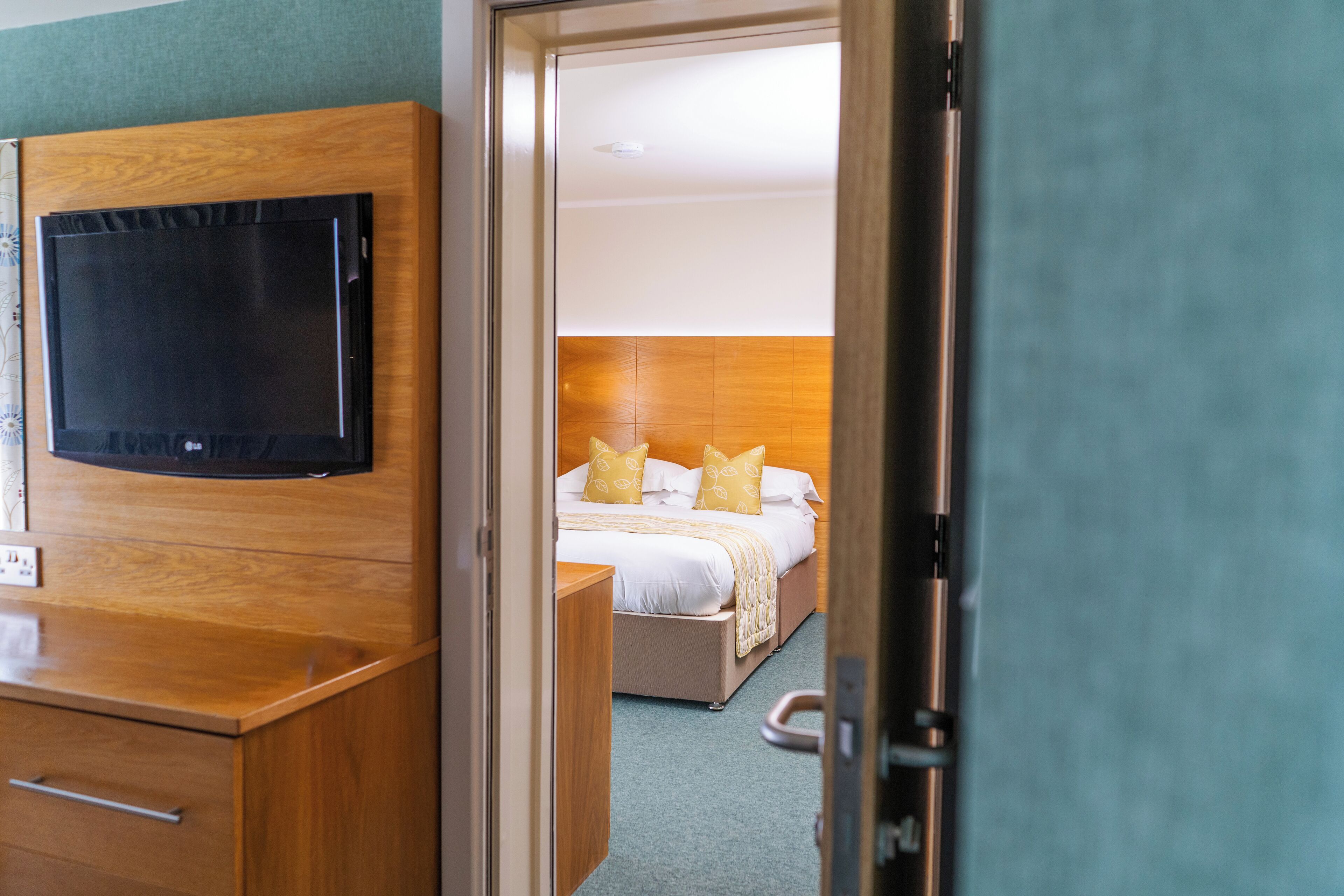 Hotellihuone, jossa on puinen vaatekaappi, seinälle asennettu litteänäyttöinen televisio, valkoisilla lakanoiden ja keltaisilla tyynyillä varustettu sänky sekä pieni yöpöytä.