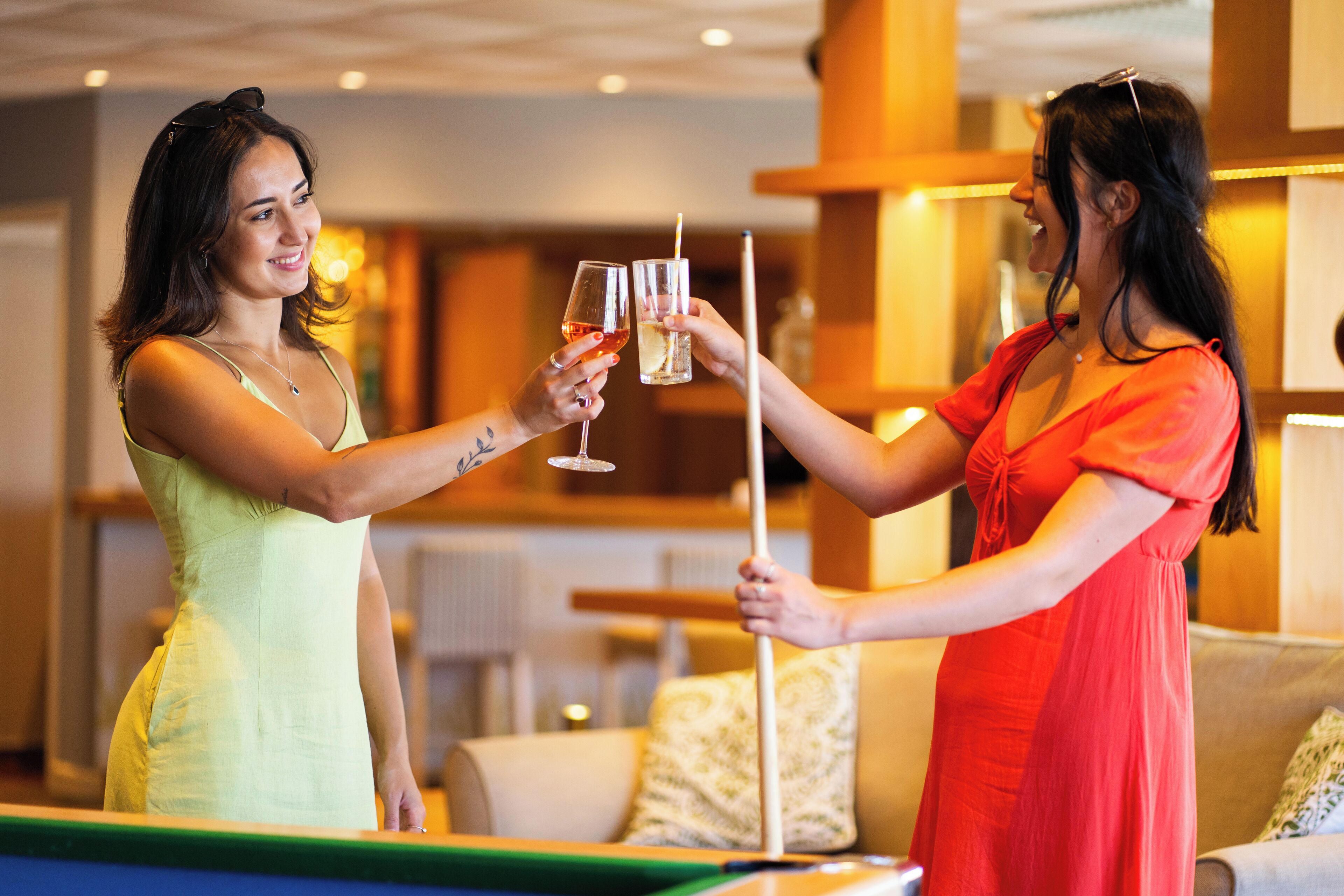 Two people toasting with drinks in a room with a pool table, a sofa, and a lamp.