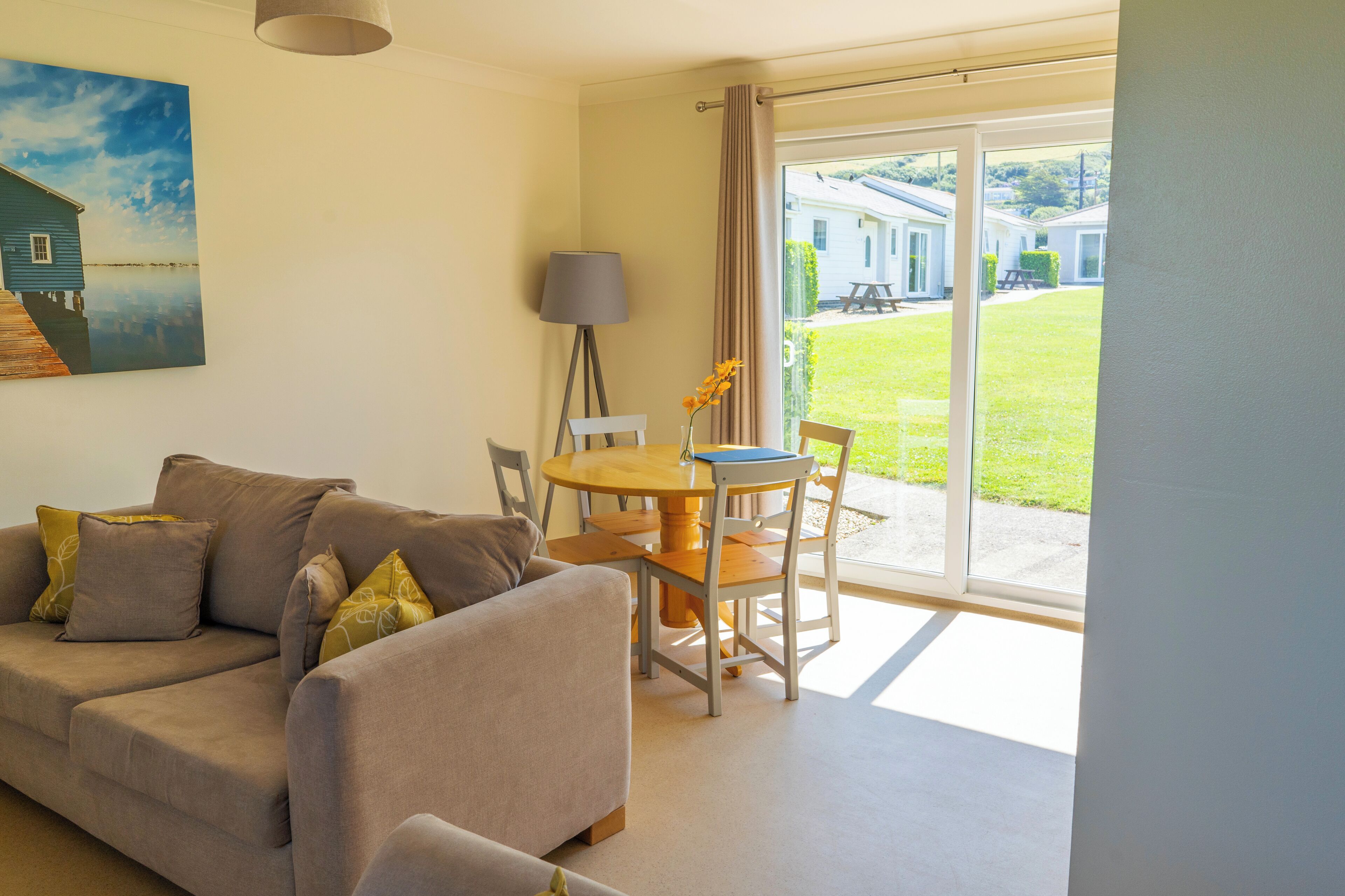 A living room with a sofa, a table, and chairs. There is a painting on the wall and a view of a house and lawn outside.