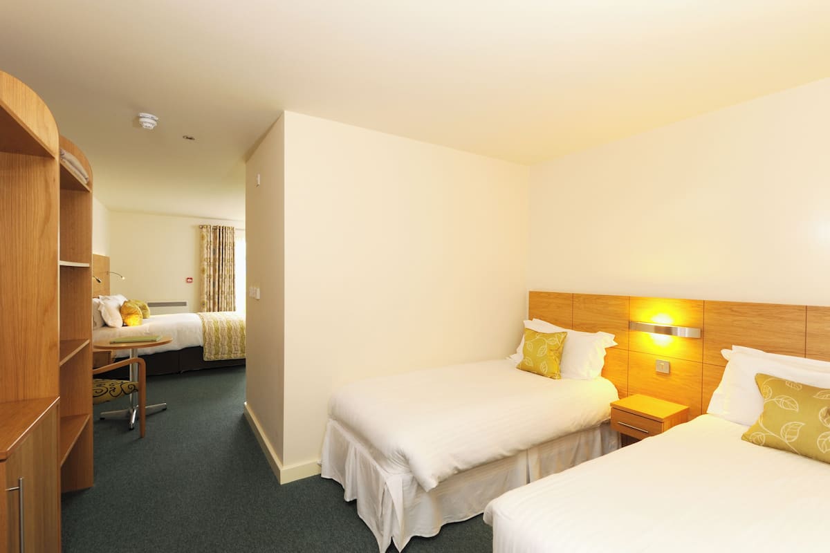 A hotel room with two beds, a wooden wardrobe, a bedside table, and a wall-mounted light fixture.