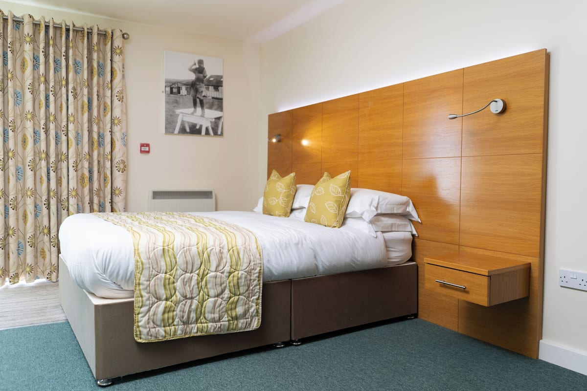 A neatly made bed with a wooden headboard, a bedside drawer, and a patterned curtain.