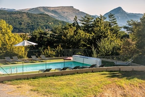 A swimming pool with lounge chairs and a covered area, surrounded by trees and mountains in the background. - Hôtel Le Panoramic (La Palud-sur-Verdon)