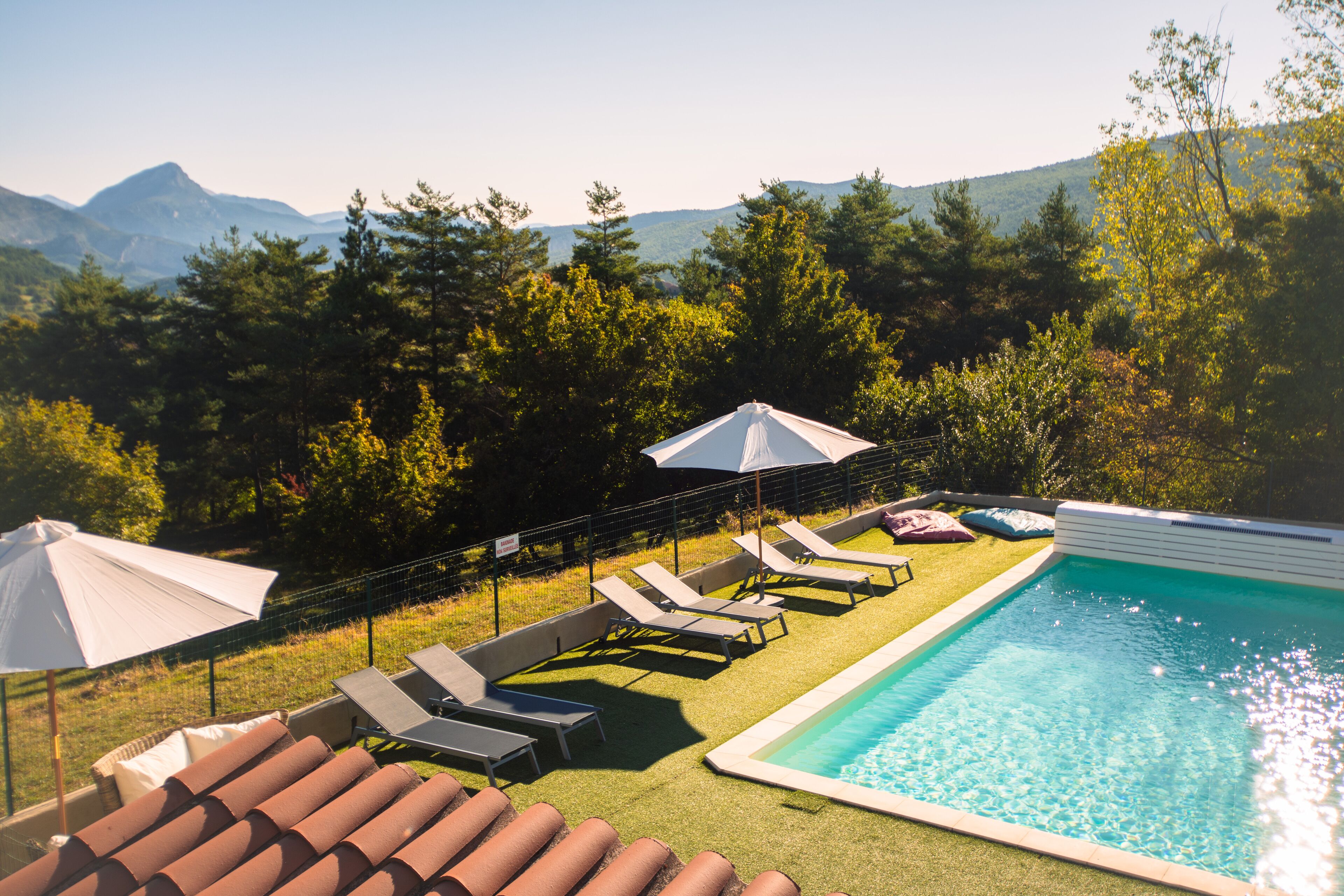 Una piscina con lettini e ombrelloni, circondata da un'area verde e alberi, con montagne sullo sfondo.