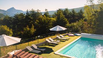 Una piscina con lettini e ombrelloni, circondata da un'area verde e alberi, con montagne sullo sfondo.