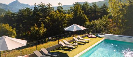 Une piscine entourée de chaises longues et de parasols, elle-même bordée d’une pelouse et d’arbres, avec des montagnes en toile de fond.
