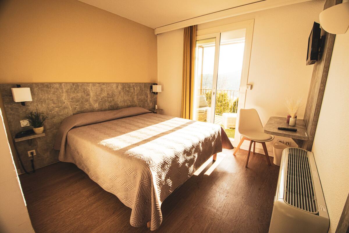 A hotel room with a bed, bedside table, lamp, chair, and a view of the outdoors.