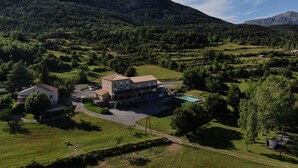 A large building with multiple rooms, a swimming pool, and a view of the surrounding green landscape and mountains. - Hôtel Le Panoramic (La Palud-sur-Verdon)