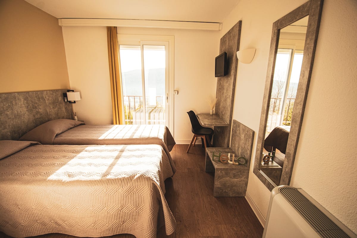 A hotel room with two beds, a desk, a chair, a mirror, and a view of the outdoors.