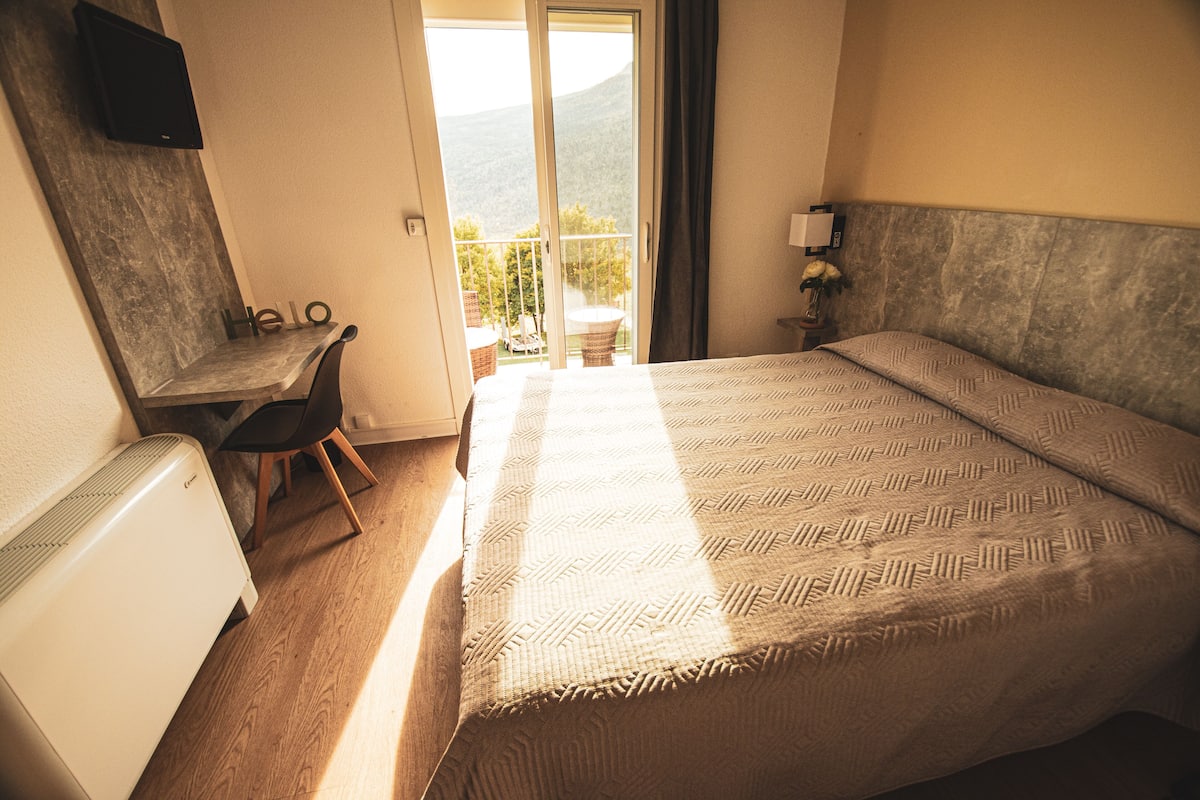 A hotel room with a bed, a desk, a chair, a TV, and a view of the outdoors.