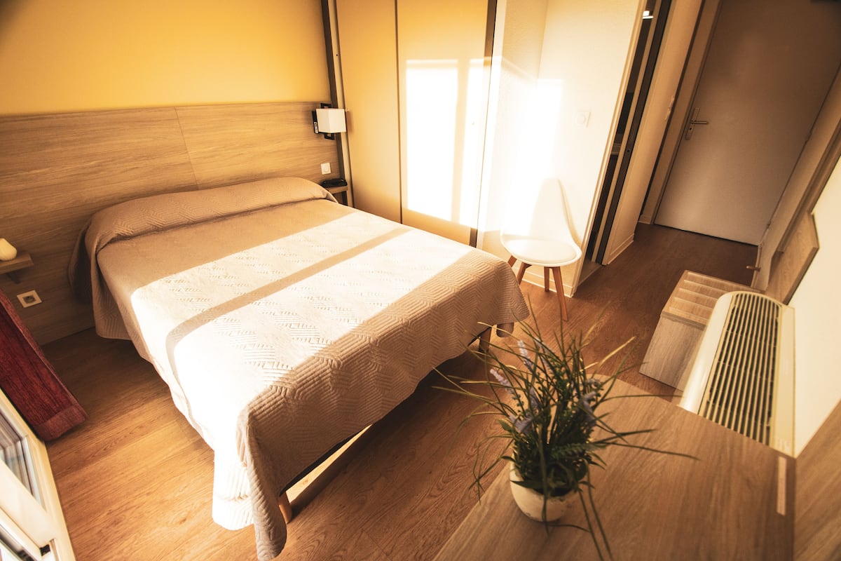 A modern hotel room with a wooden floor, a large bed with a beige bedspread, a bedside table with a lamp, a potted plant, and a radiator.