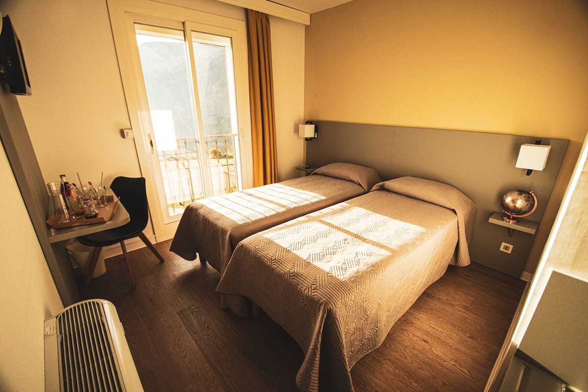 A hotel room with two beds, a desk with a chair, a window with curtains, and a small table with a globe and lamp.