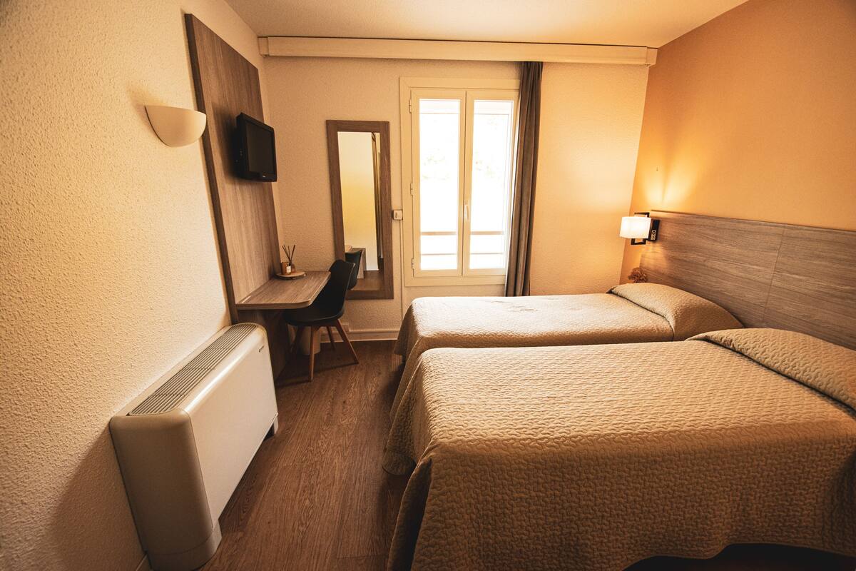 A hotel room with two beds, a desk, a chair, a television, and a window with curtains.