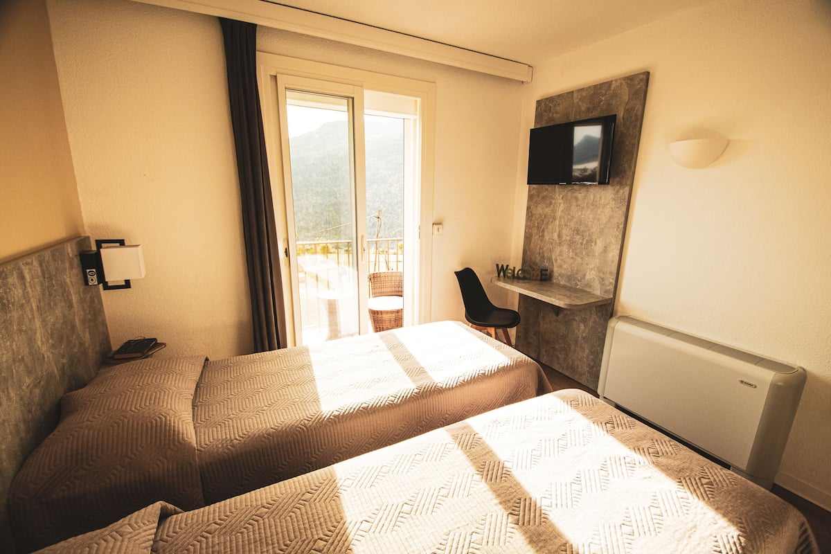 A hotel room with two beds, a TV mounted on the wall, a desk with a chair, and a view of the outdoors through a sliding door.