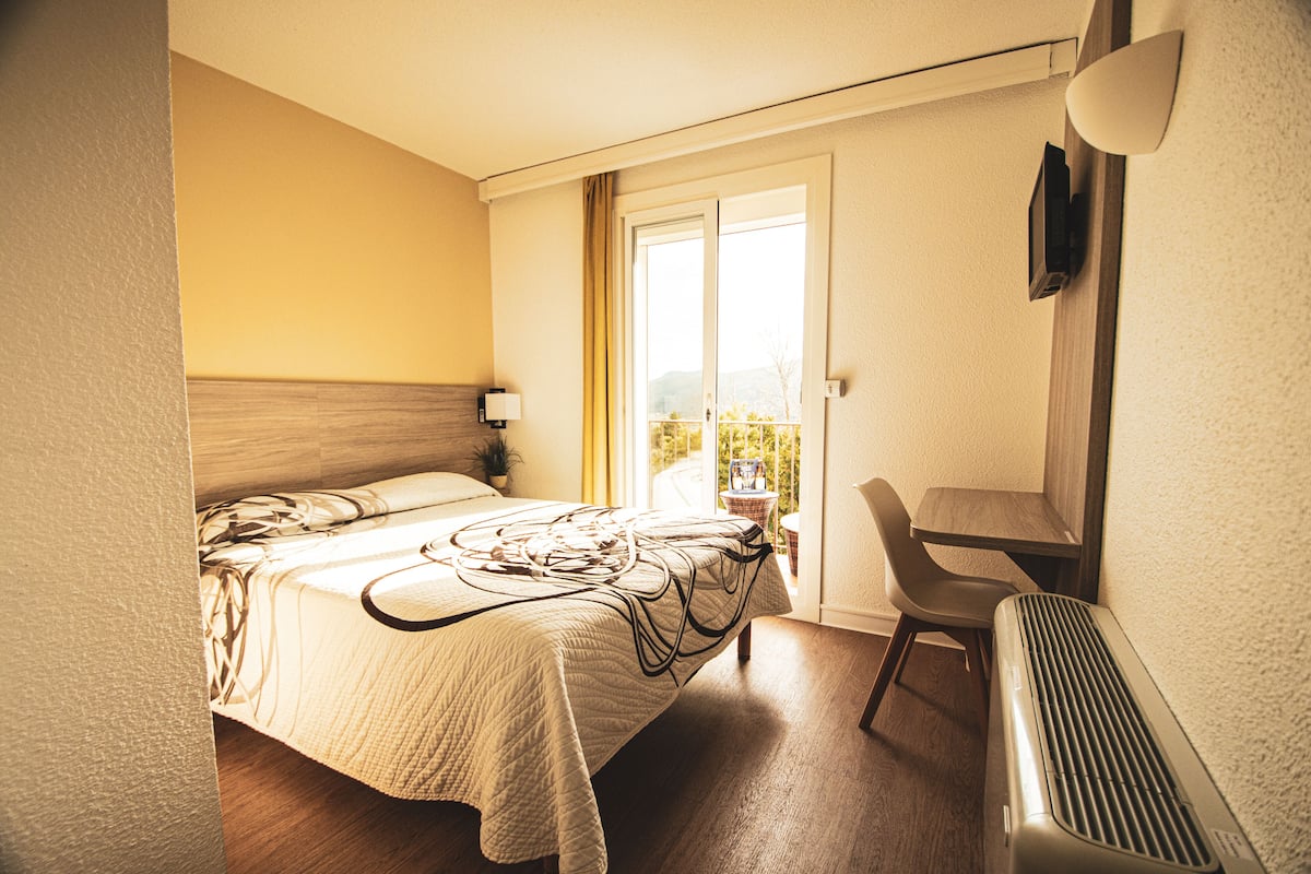 A hotel room with a bed, a chair, a desk, and a radiator.
