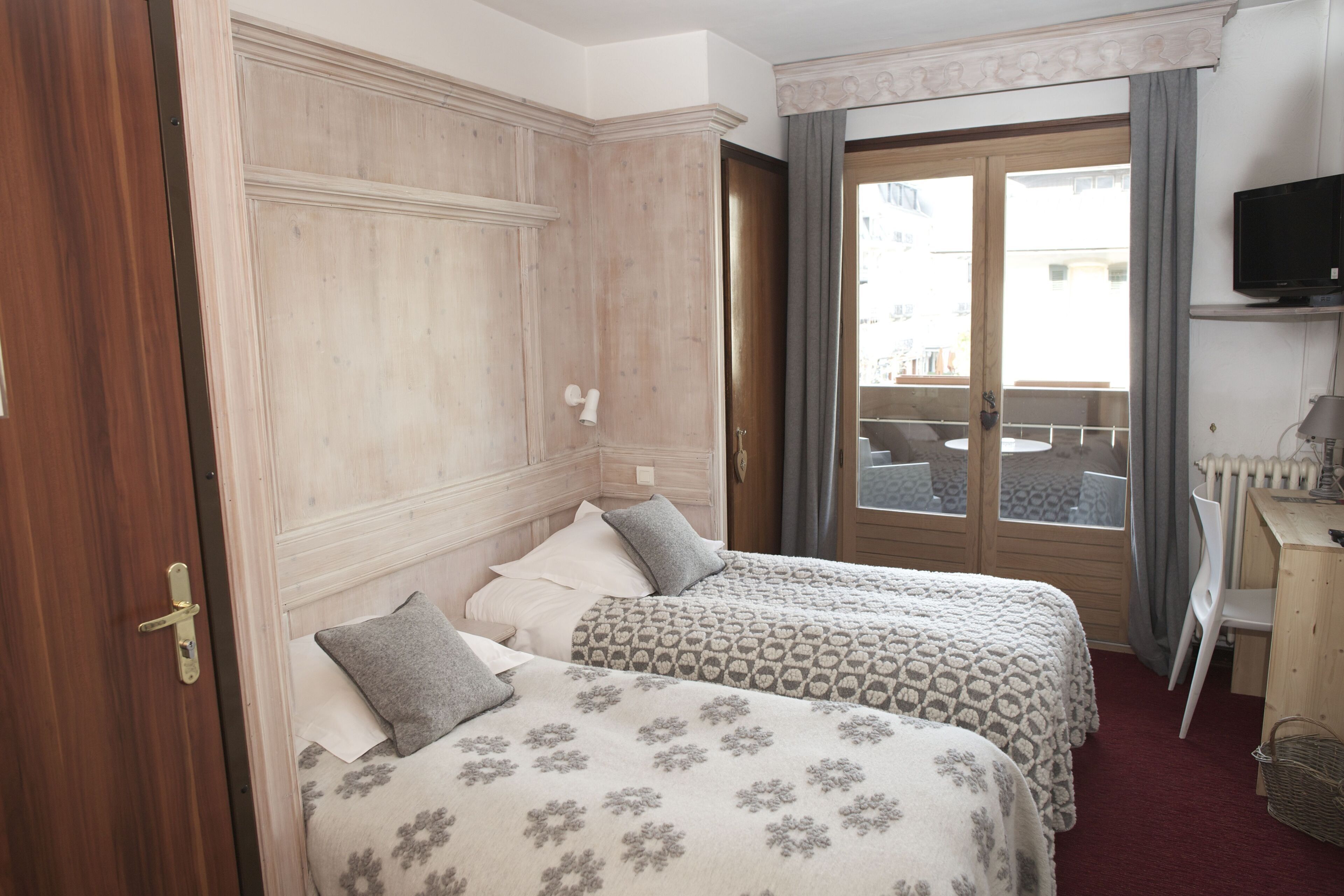 A hotel room with two beds, a wooden wardrobe, a desk with a chair, a television, and a balcony with a view.
