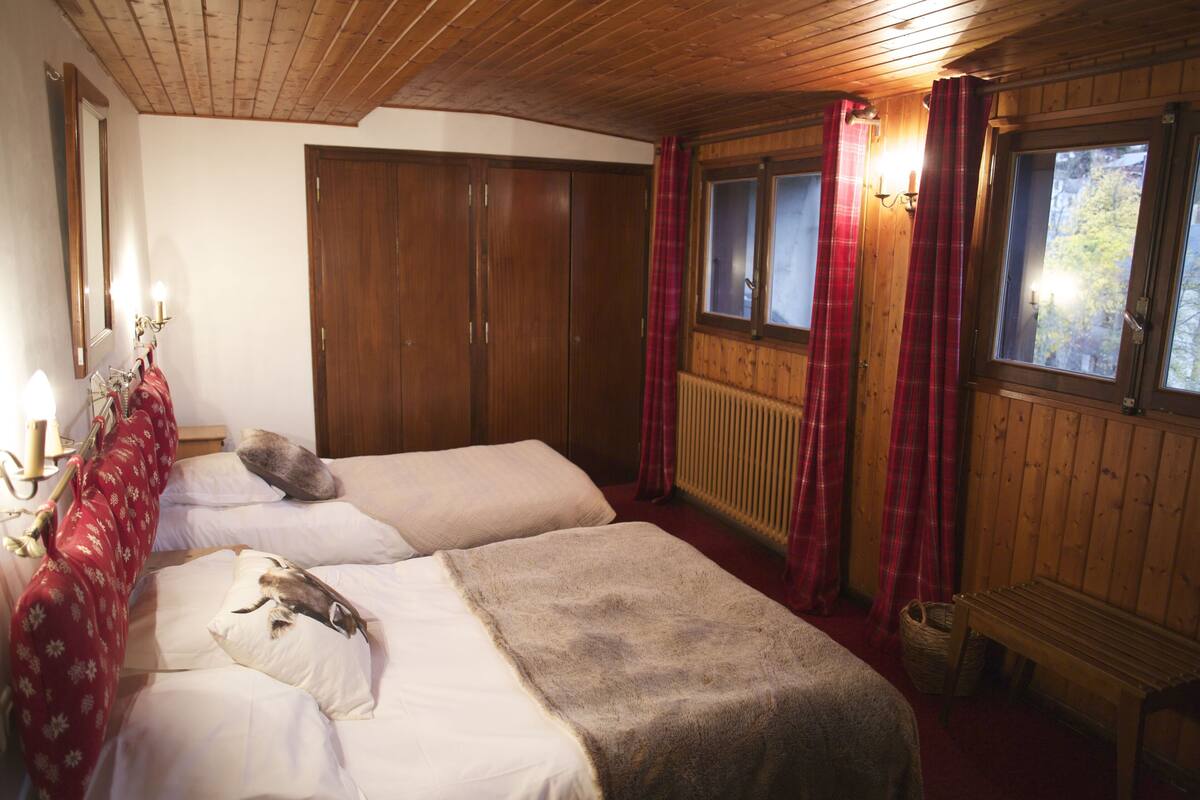Una habitación con dos camas, armario de madera, cortinas rojas y ventana con vistas a árboles.