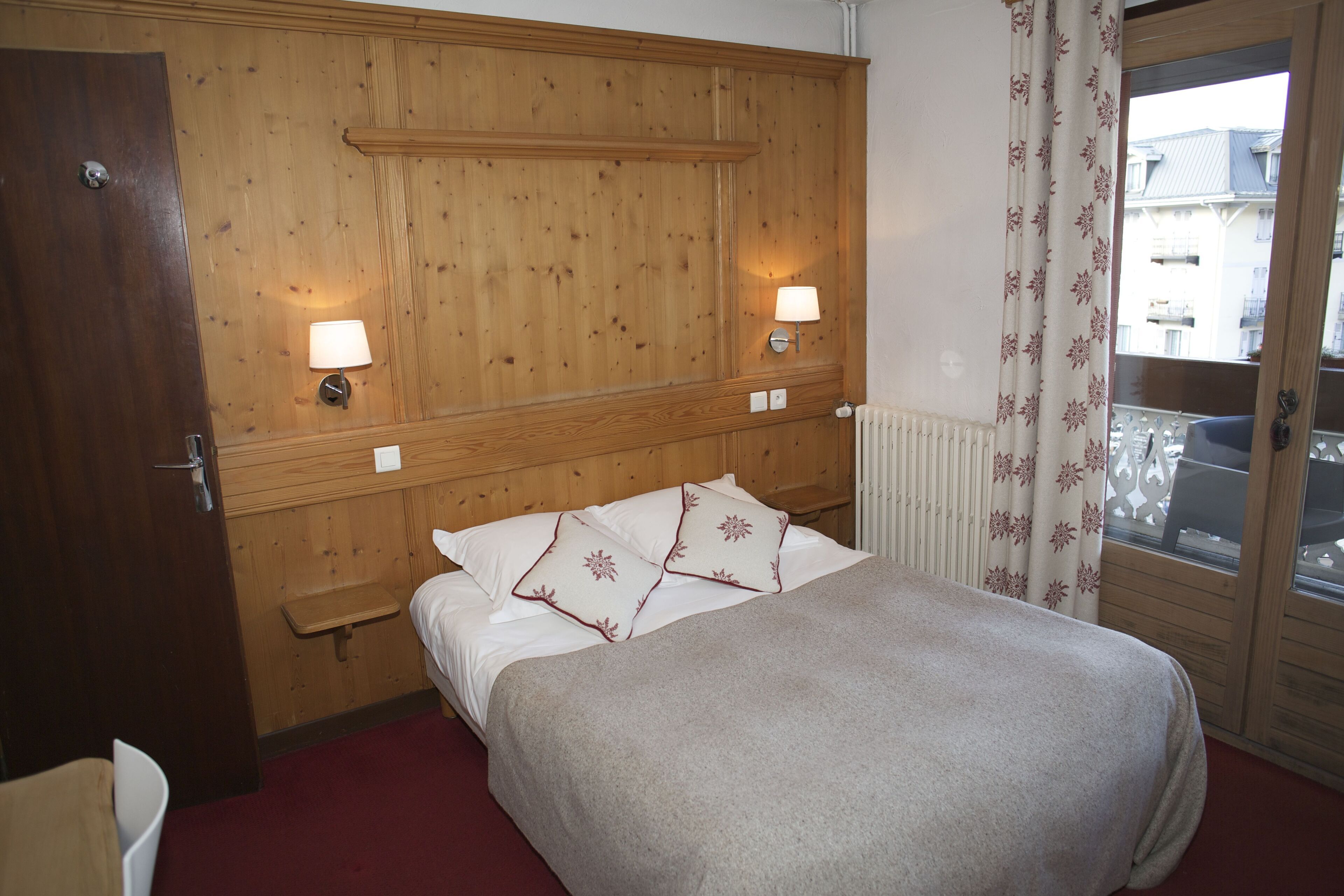A hotel room with a wooden headboard, two bedside lamps, a bed with white bedding and decorative pillows, a small bedside table, and a view through a window with curtains.