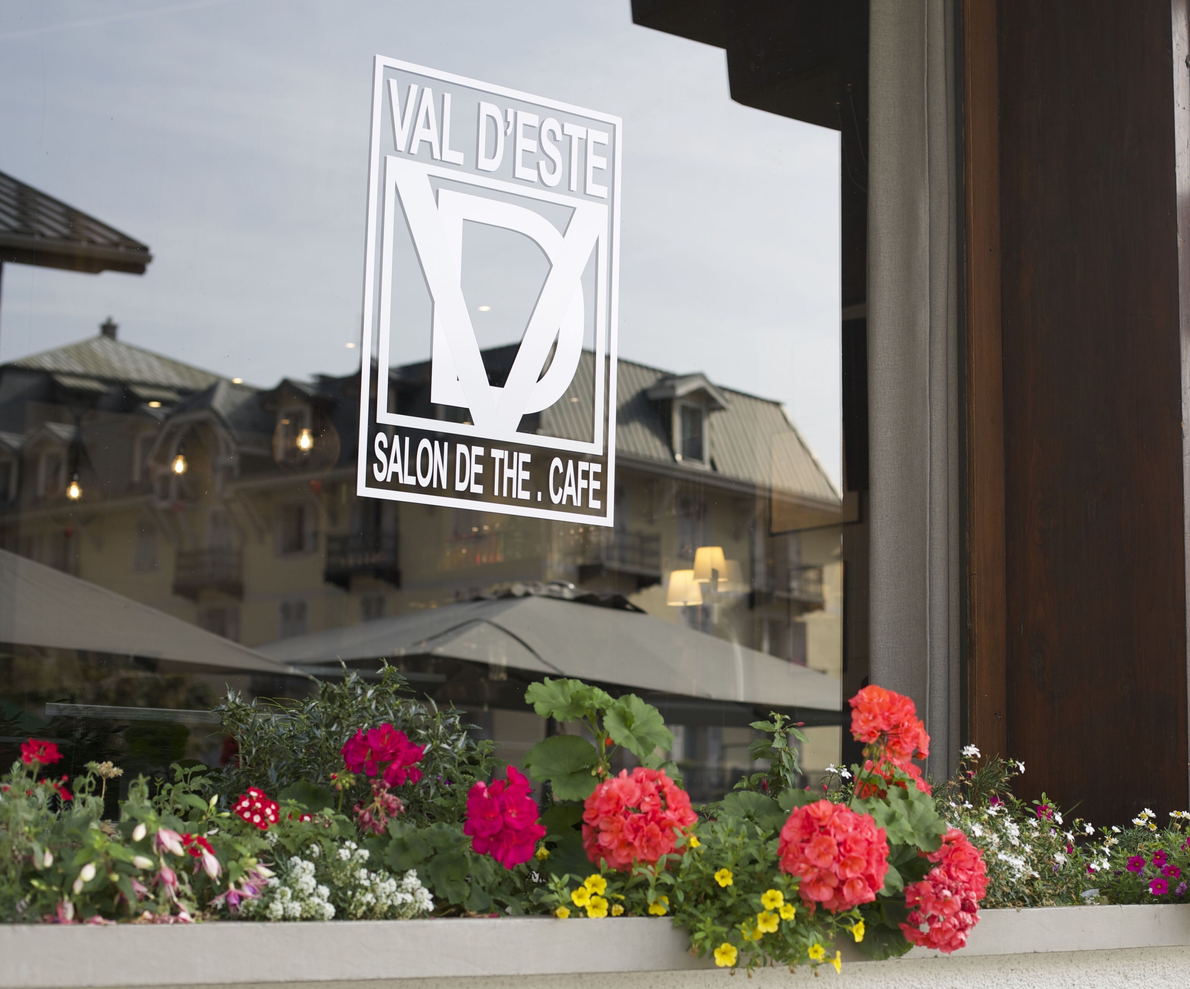 Schild für "VAL D'ESTE SALON DE THE . CAFE" mit Blick von einem Balkon auf bunte Blumen und ein Gebäude im Hintergrund.