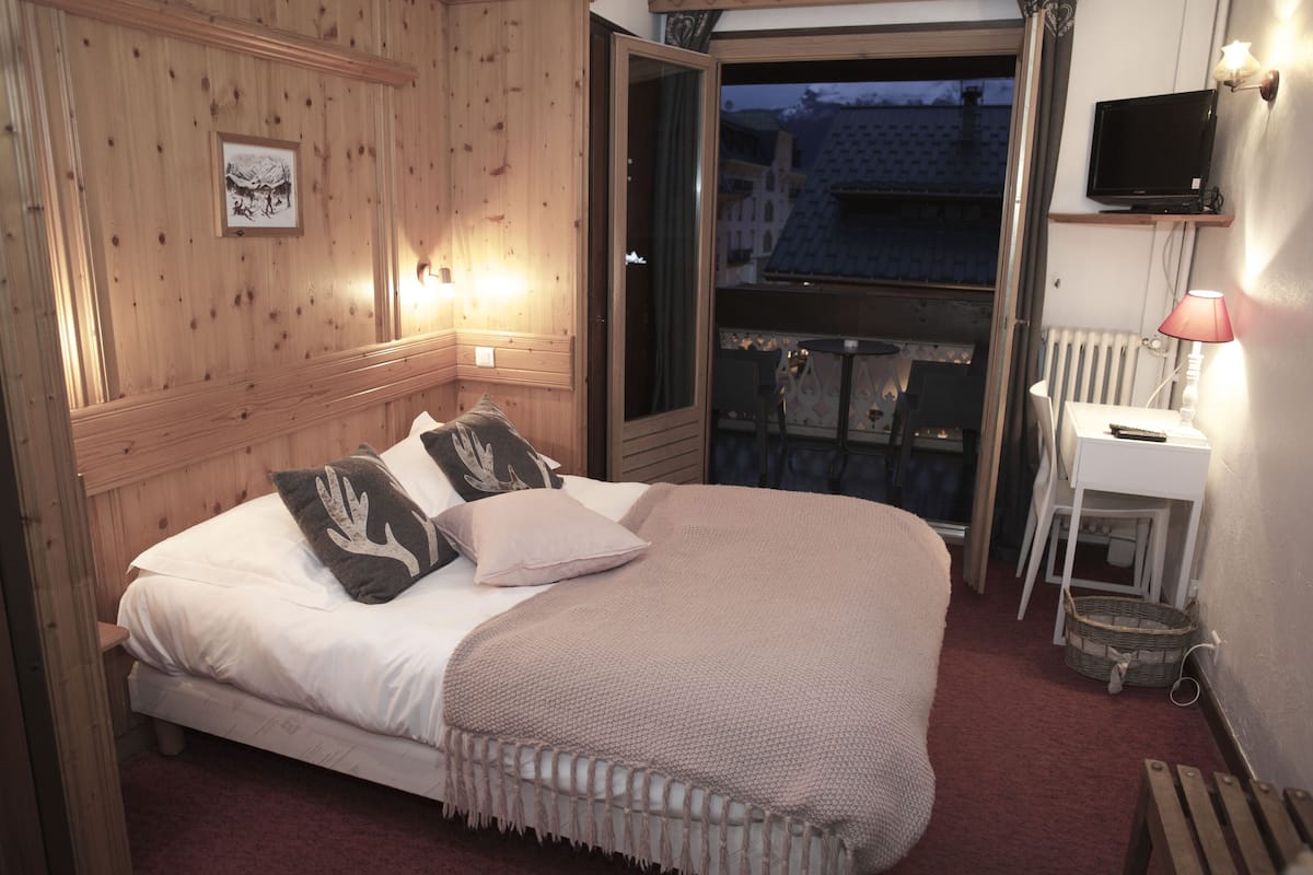Un dormitorio con cama, almohadas, una mesita de noche, lámpara, televisor y ventana con vistas a edificios.