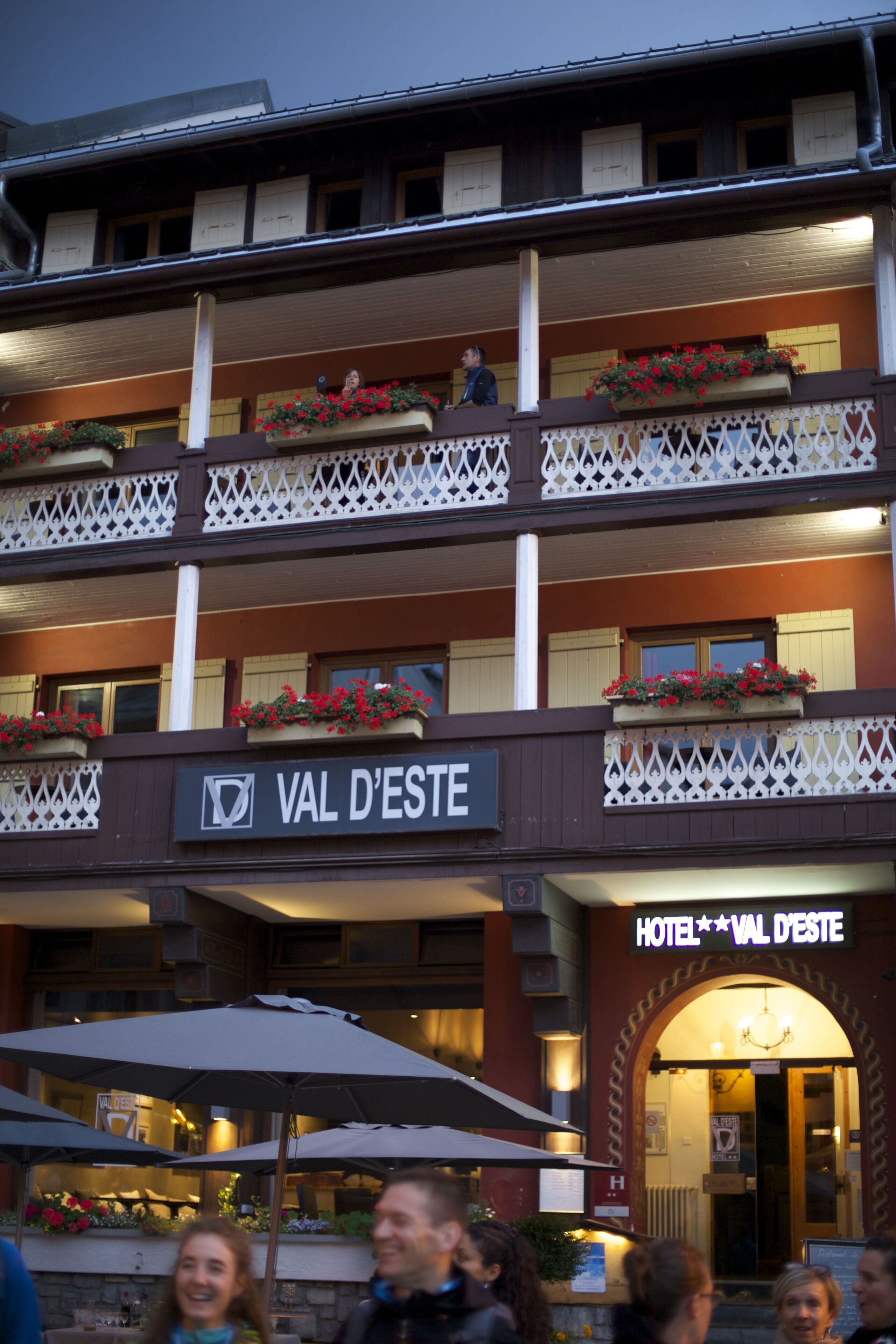 A hotel with balconies adorned with flower boxes, a sign reading "VAL D'ESTE," and outdoor seating with umbrellas.