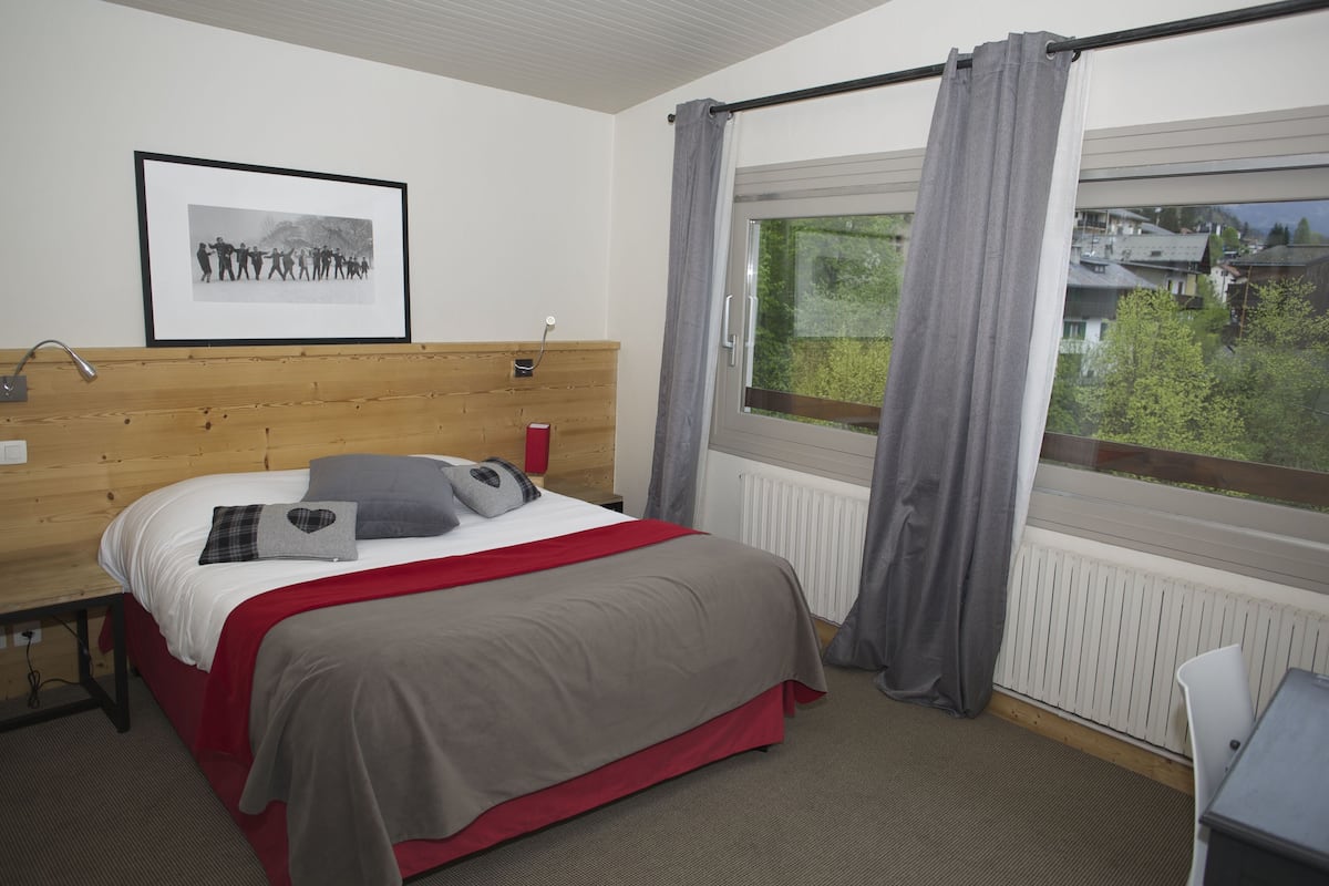 Habitación de hotel con cabecera de madera, cama con ropa de cama roja y gris, fotografía blanco y negro enmarcada en la pared, ventana con cortinas grises y vistas a zonas verdes.