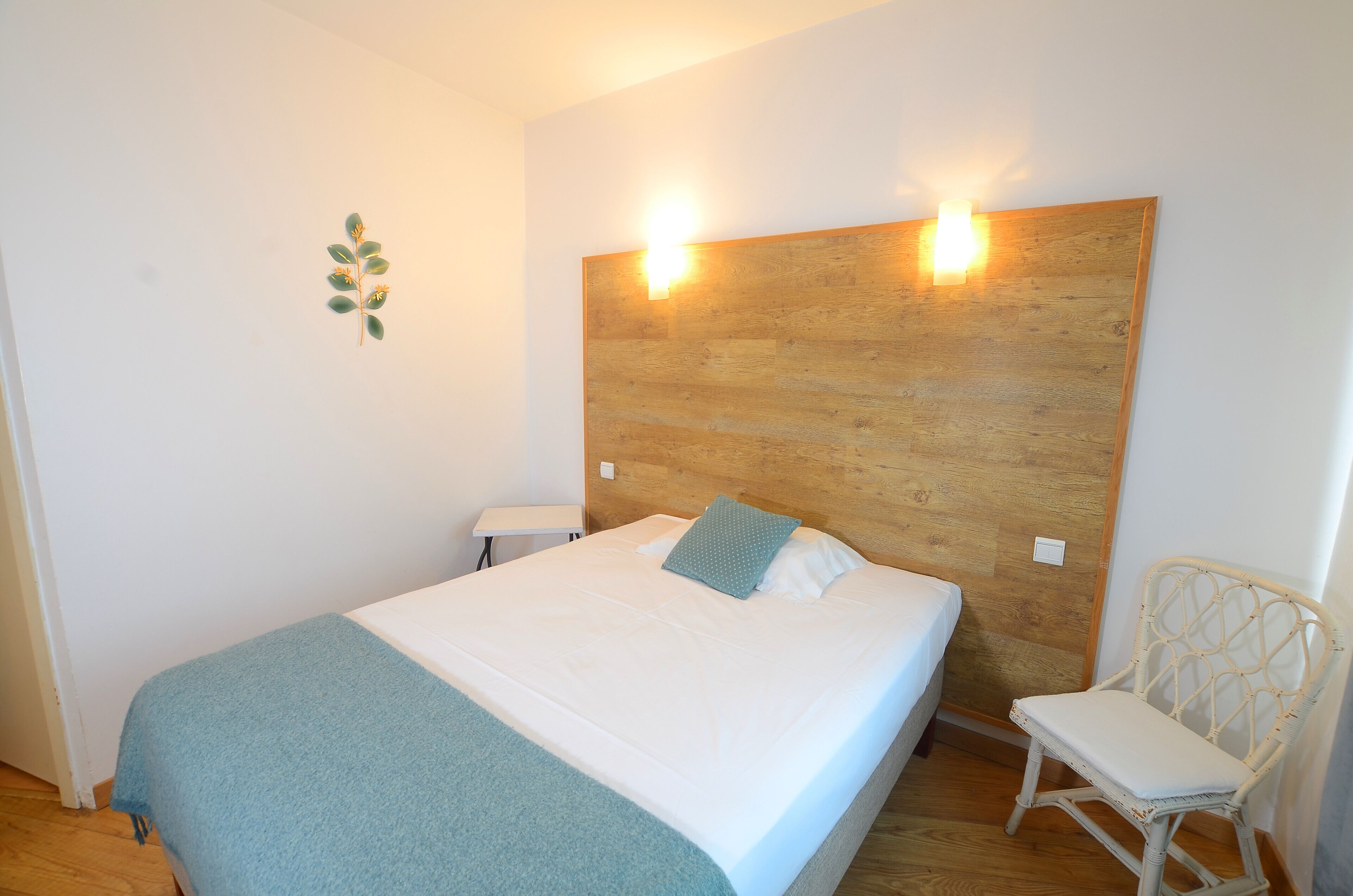 A bedroom with a wooden headboard, a bed with white bedding, a blue throw blanket, a white chair, and a small table.
