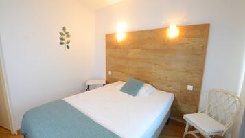 Un dormitorio con cabecera de madera, cama con ropa de cama blanca, manta azul, una silla blanca y una mesita.