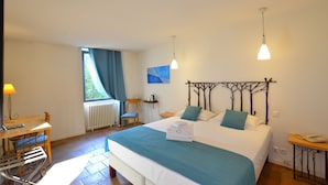 A hotel room with a large bed, a desk with a lamp, a chair, a window with curtains, and a painting on the wall.