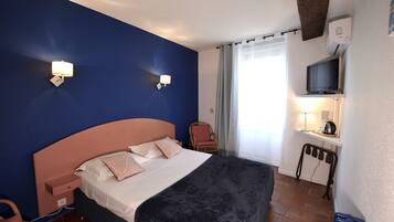 Habitación de hotel con cama, mesita de noche, lámpara, silla, televisión y una pequeña mesa con lámpara.
