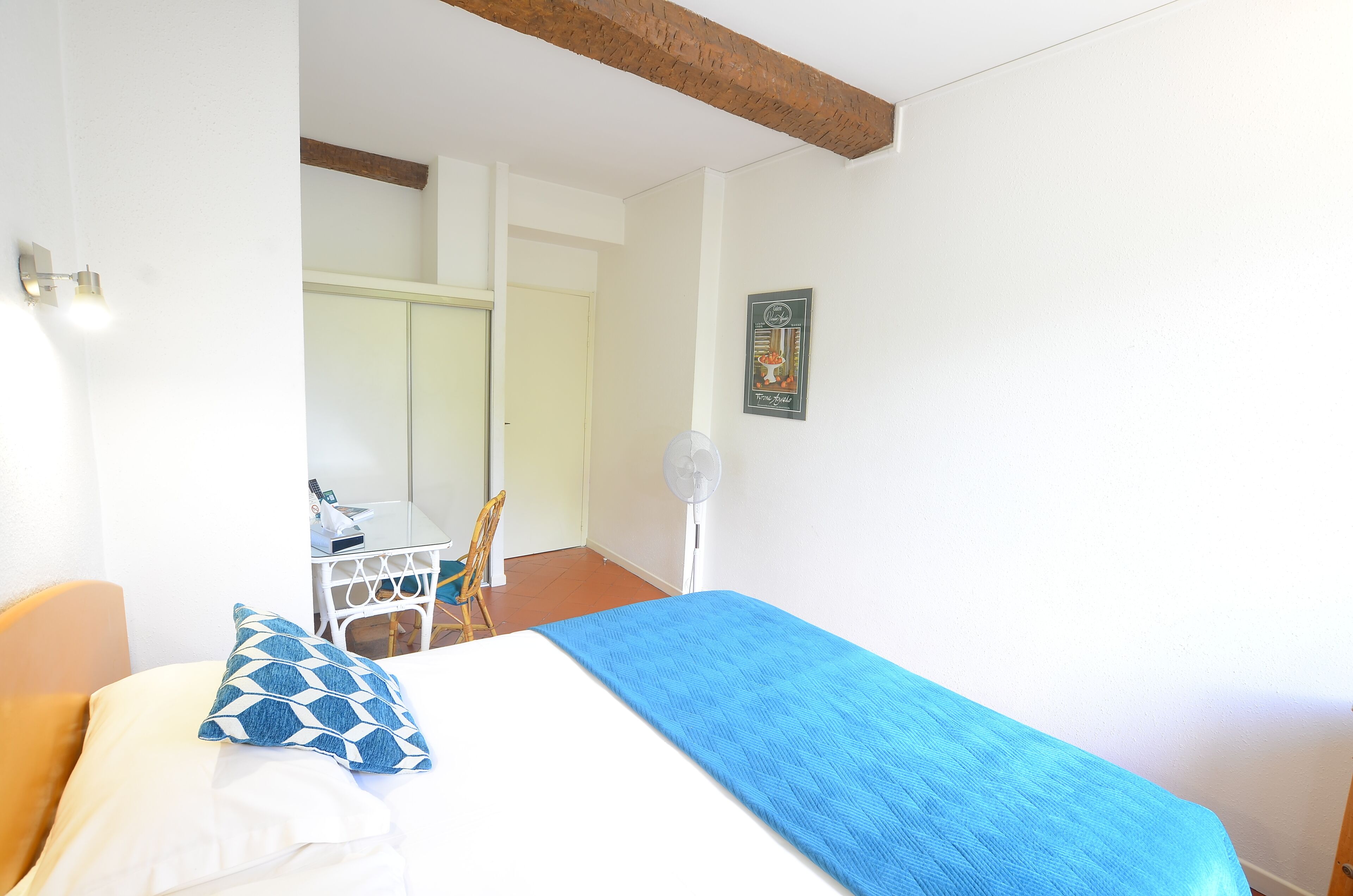 A room with a bed, a chair, a table, and a wall-mounted fan.