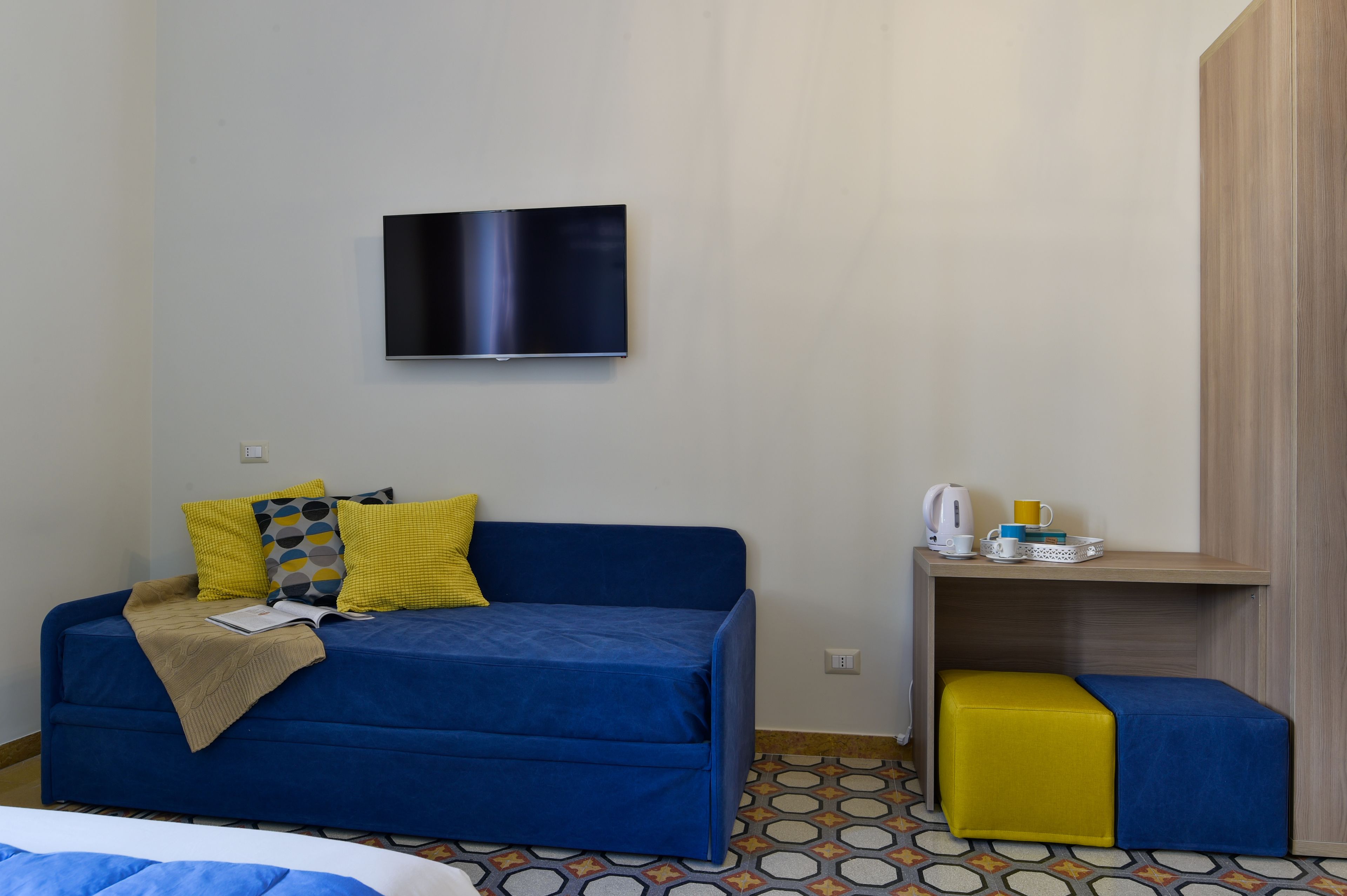 A room with a blue sofa, a yellow ottoman, a TV mounted on the wall, and a small table with a kettle and cups.