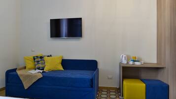 A room with a blue sofa, a yellow ottoman, a TV mounted on the wall, and a small table with a kettle and cups.