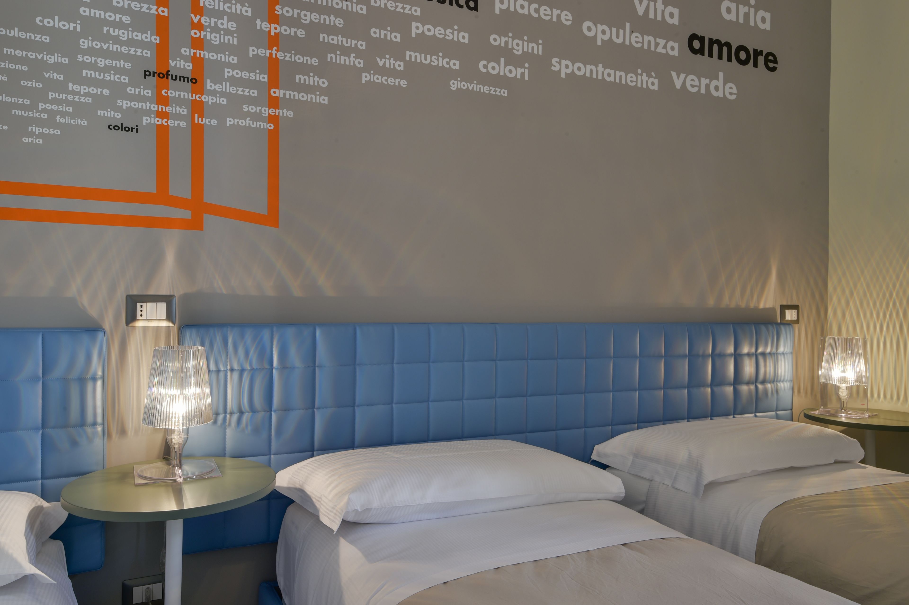 Una habitación de hotel con cabecera azul, dos camas y una pared con texto en italiano.