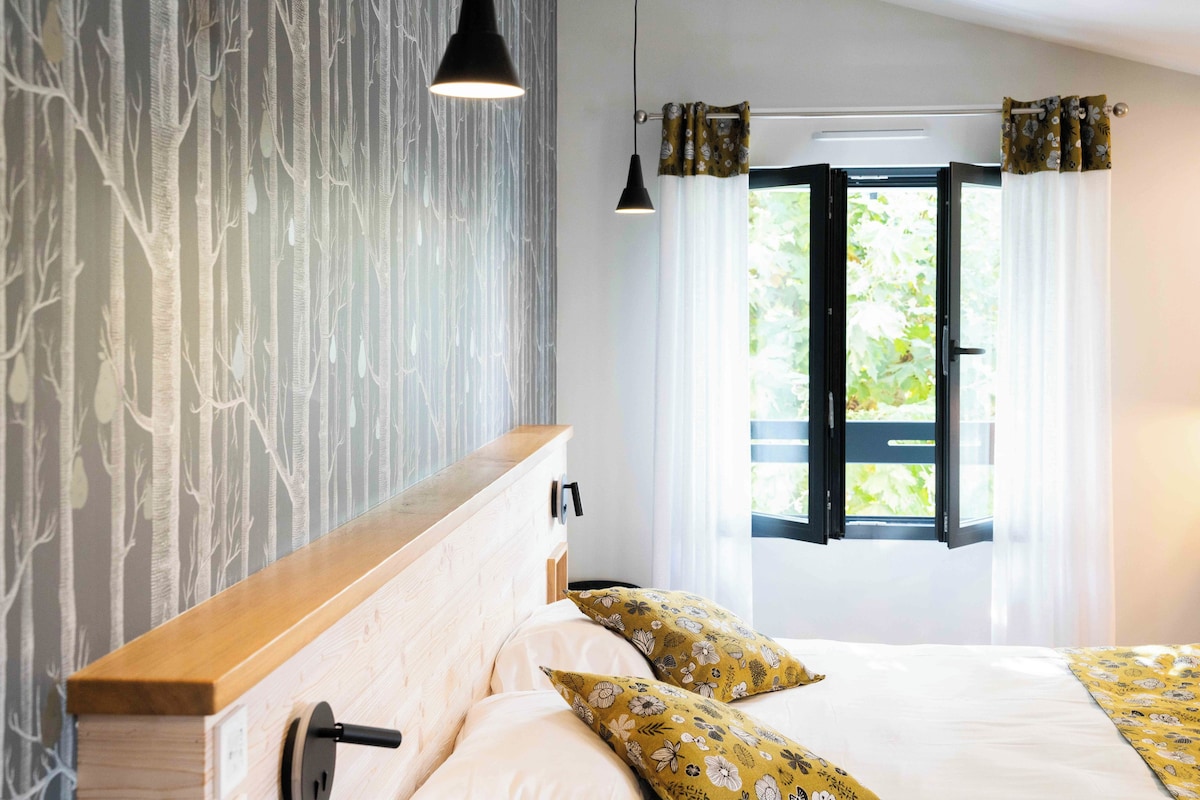 Een slaapkamer met een houten hoofdbord, een bed met bloemenkussens, een raam met gordijnen en een muur met een boommotief.