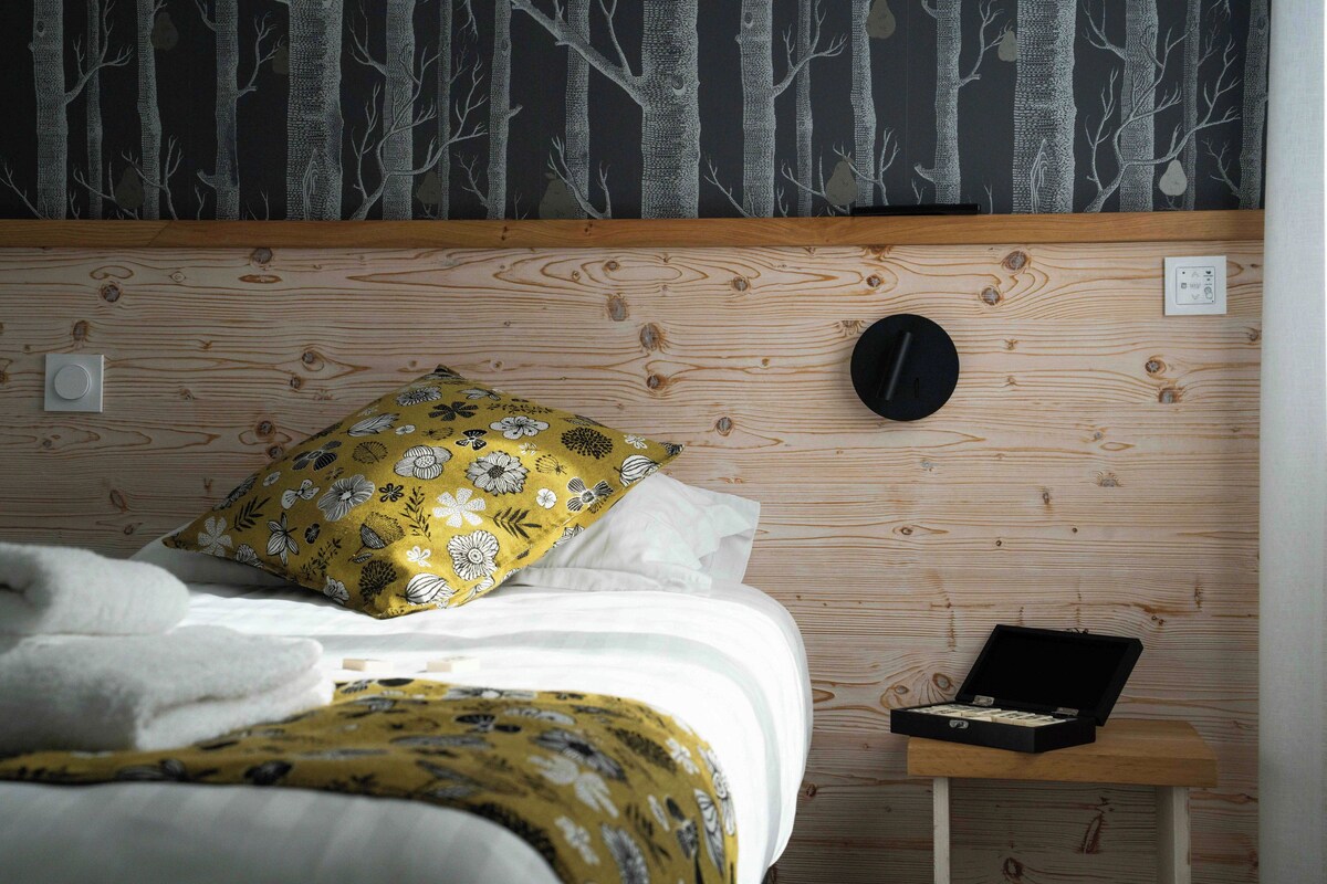 Un dormitorio con cabecera de madera, cama con ropa de cama blanca, almohada floral amarilla, mesita de noche con un objeto negro y pared con estampado de bosque.
