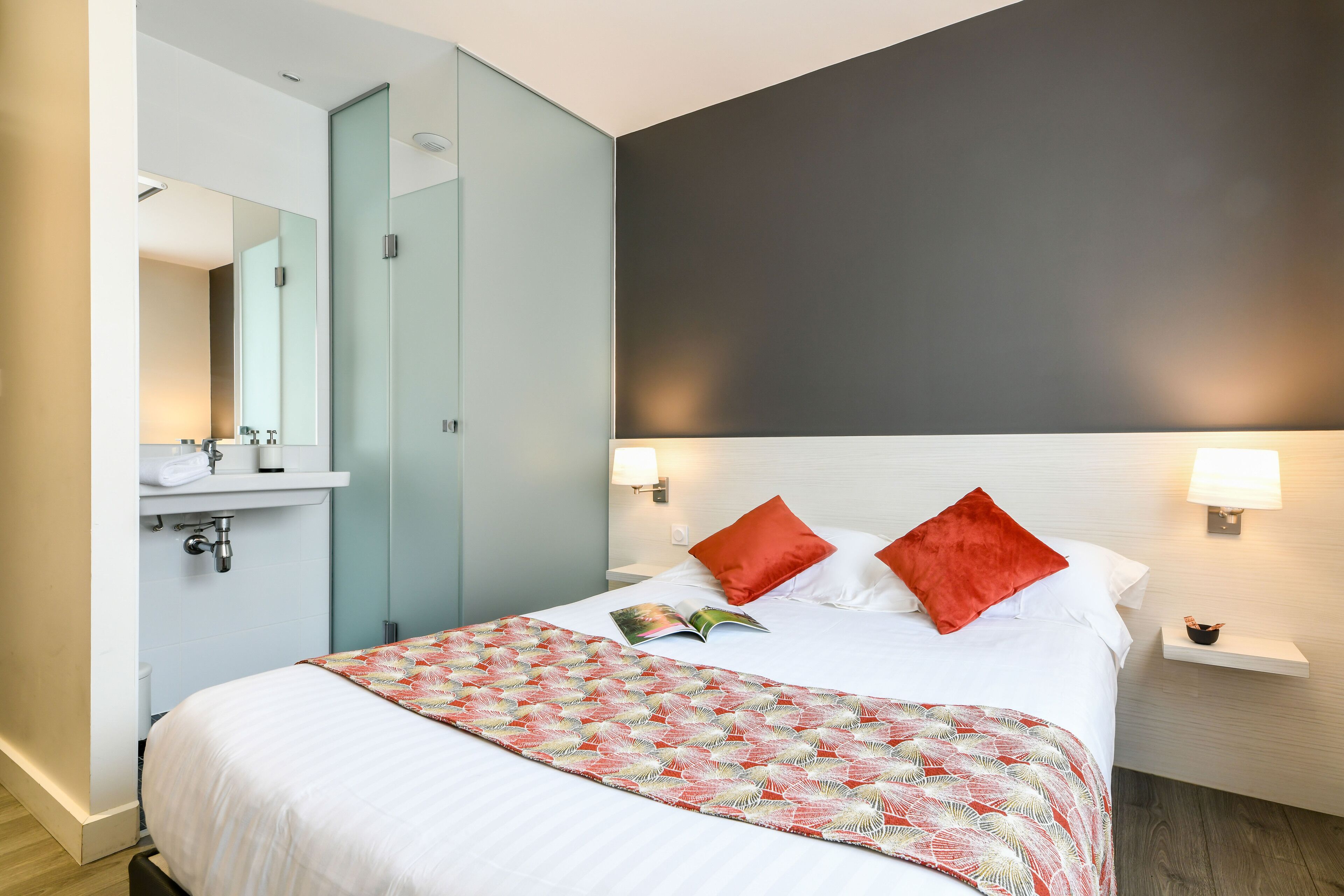 Habitación de hotel con cama, mesita de noche, espejo, lavabo y una manta roja.