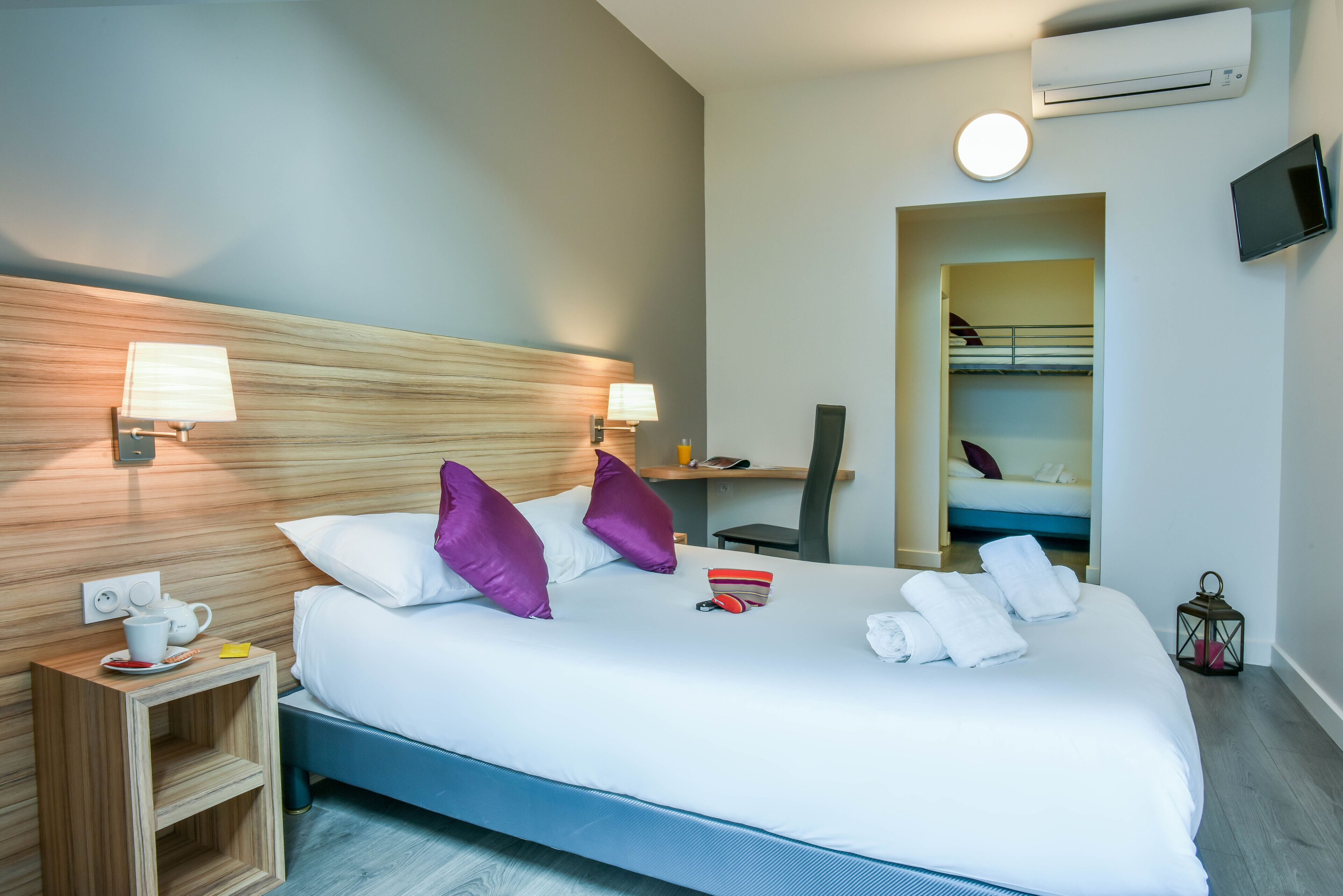 A hotel room with a large bed, two bedside lamps, a desk, a chair, a TV, and an air conditioning unit.