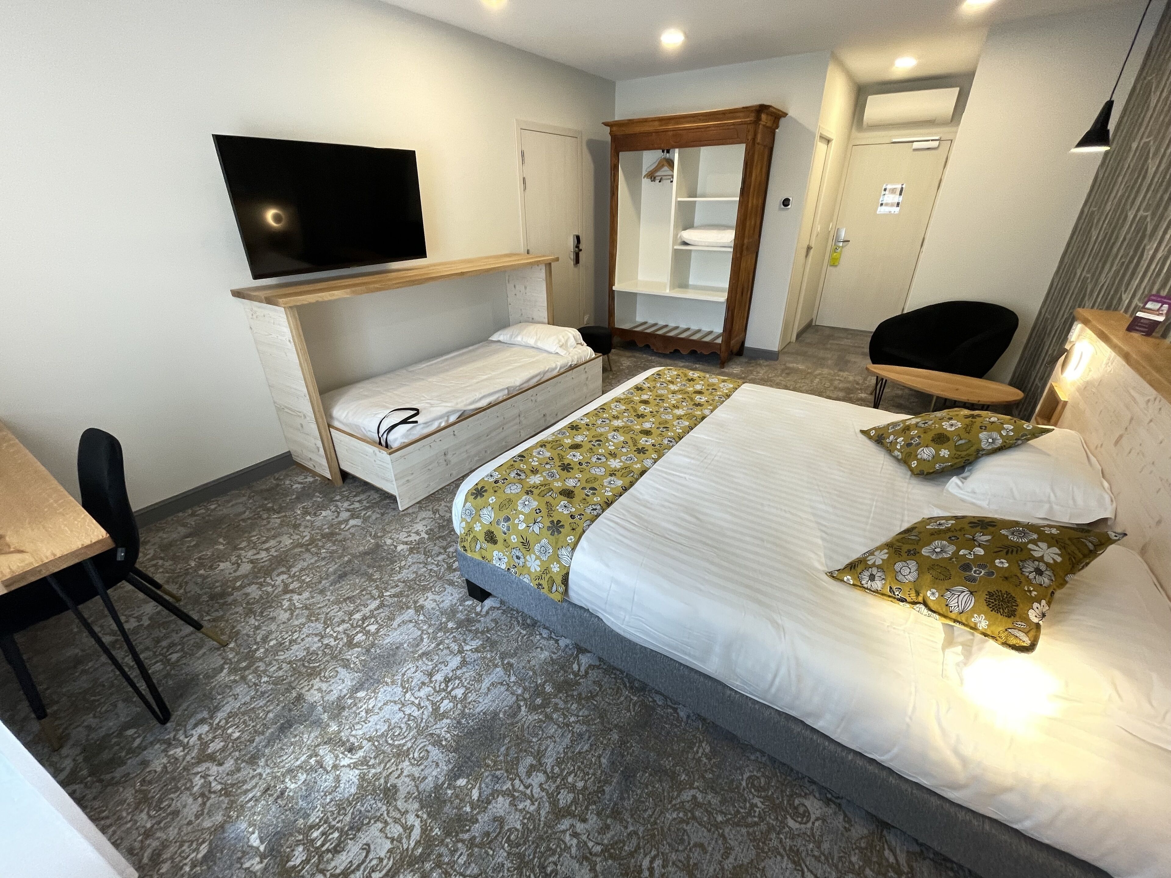 A hotel room with a bed, a TV mounted on the wall, a wooden wardrobe, a desk with a chair, and a mirror.