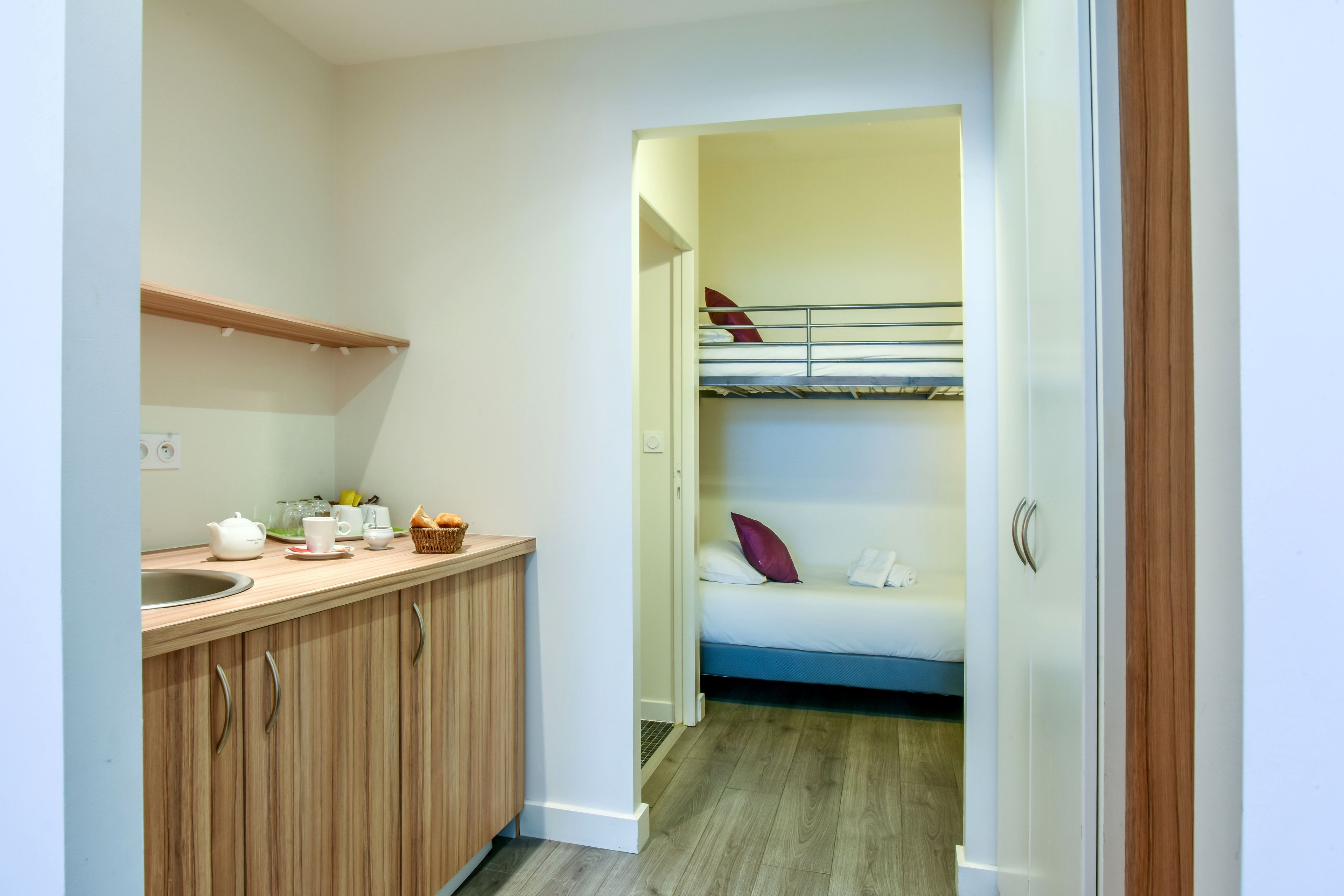 Une cuisine compacte avec un plan de travail en bois, un évier et un petit coin pour prendre le petit-déjeuner. On aperçoit un lit superposé avec des oreillers dans la pièce adjacente.