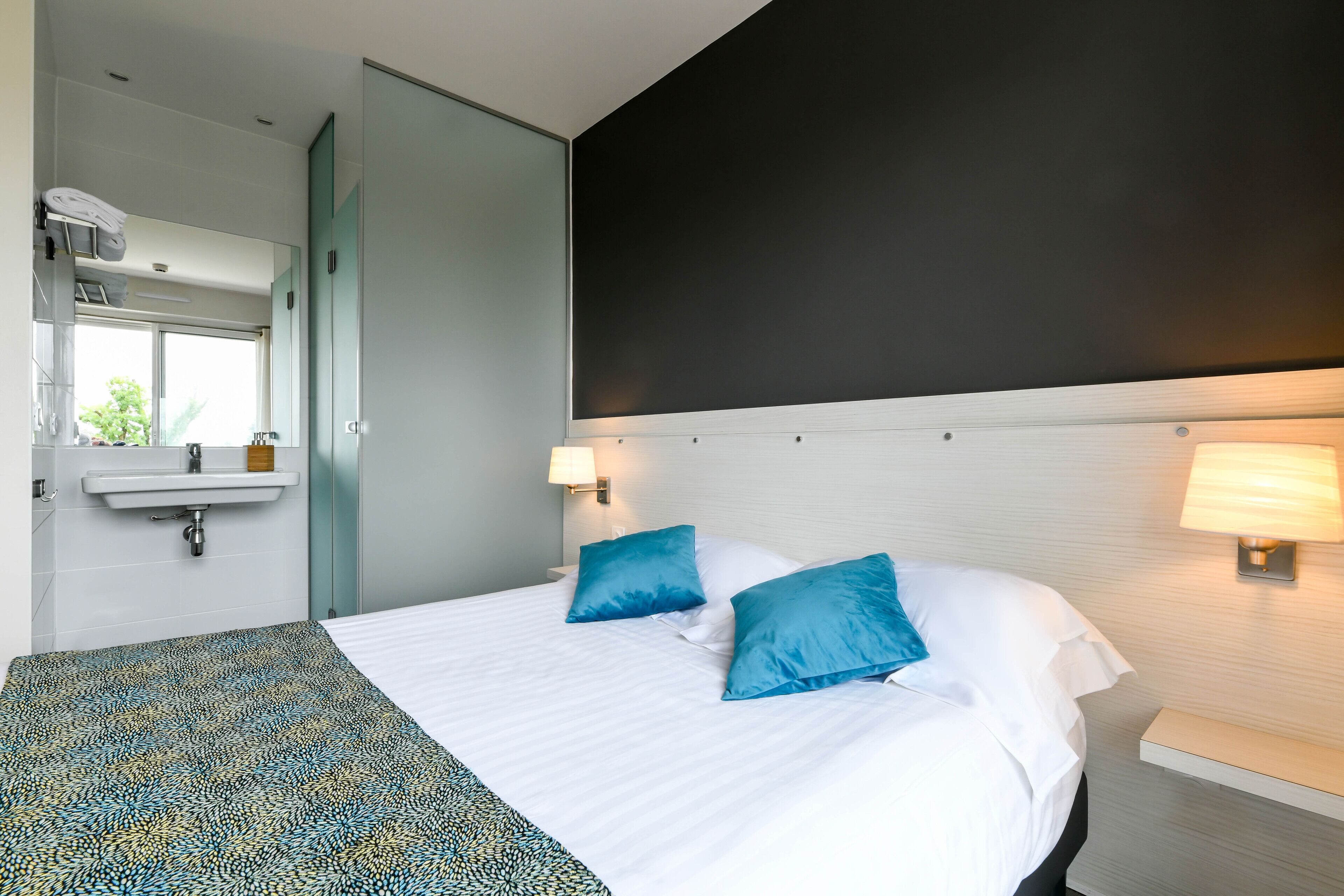 A hotel room with a bed, two pillows, a bedside table with a lamp, a mirror, a window, and a sink.