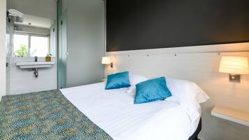 A hotel room with a bed, two pillows, a bedside table with a lamp, a mirror, a window, and a sink.