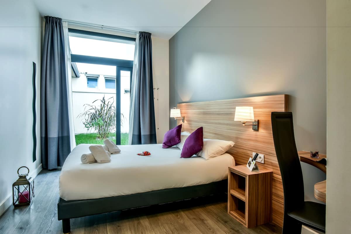 Une chambre d’hôtel moderne avec un grand lit, une tête de lit en bois, une table de chevet avec une lampe et une vue sur l’extérieur.