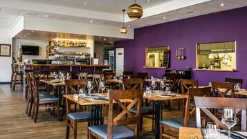 Restaurante com mesas e cadeiras de madeira, um espaço de bar com prateleiras cheias de garrafas e paredes roxas.