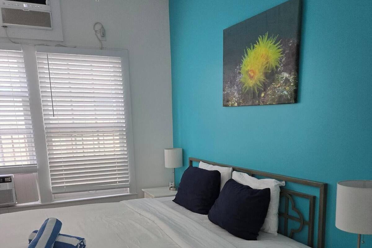 A neatly made bed with white linens and two dark pillows, a blue and white striped towel, a wall-mounted artwork of a flower, and a white bedside table with a lamp and remote control.
