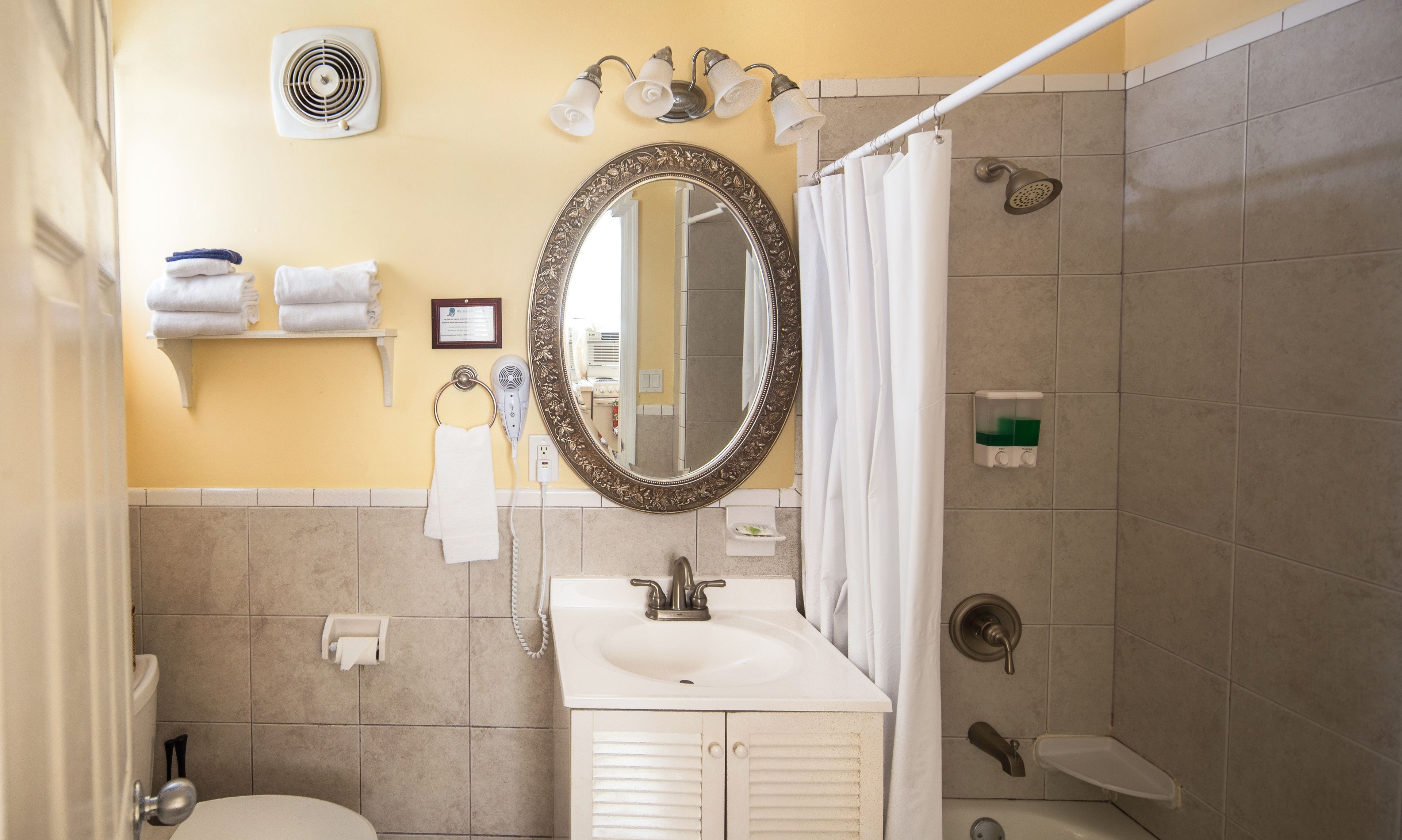 A bathroom with a sink, mirror, towel rack, shower, and a fan on the wall.