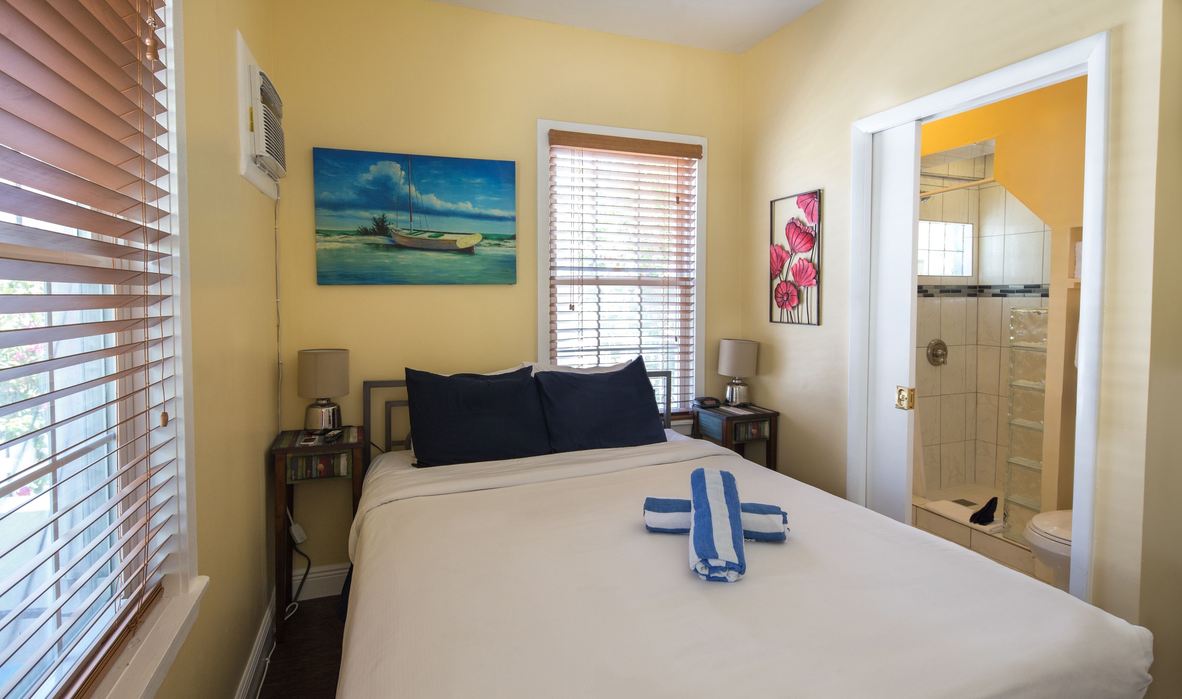 Une chambre d’hôtel avec un lit, deux tables de chevet, un tableau, une fenêtre avec des stores et une salle de bain visible à travers une porte ouverte.