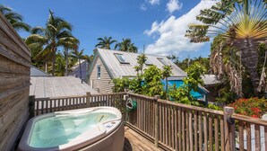 Una terraza de madera con un jacuzzi, rodeada de vegetación tropical y una casa de estilo bungalow con techo inclinado.
